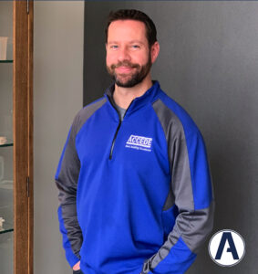 A man standing in front of a wall wearing blue and gray.